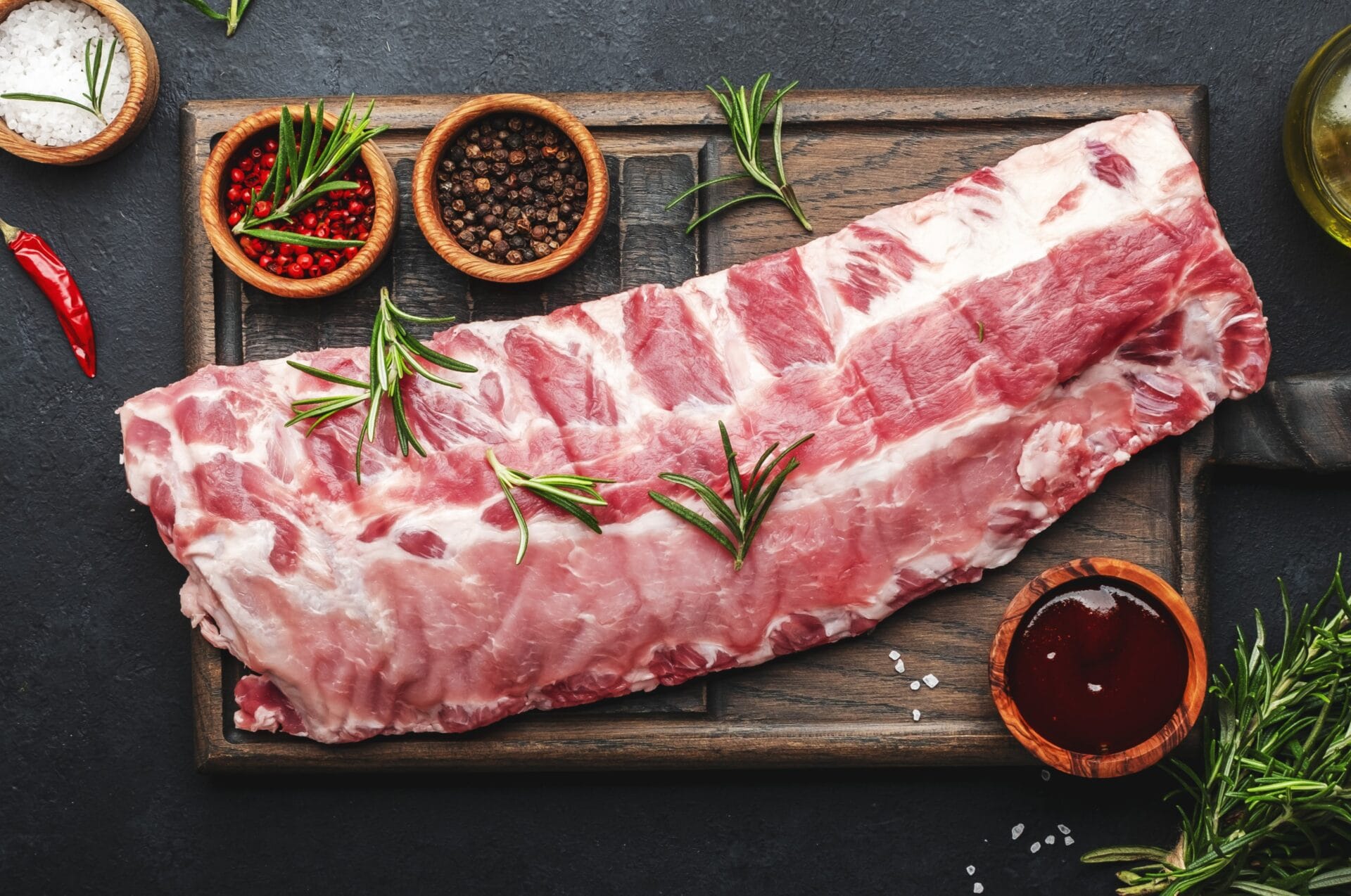 Raw Herterich's baby back pork ribs on a wooden board with spices and herbs on a dark surface