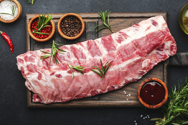 Raw Herterich's baby back pork ribs on a wooden board with spices and herbs on a dark surface