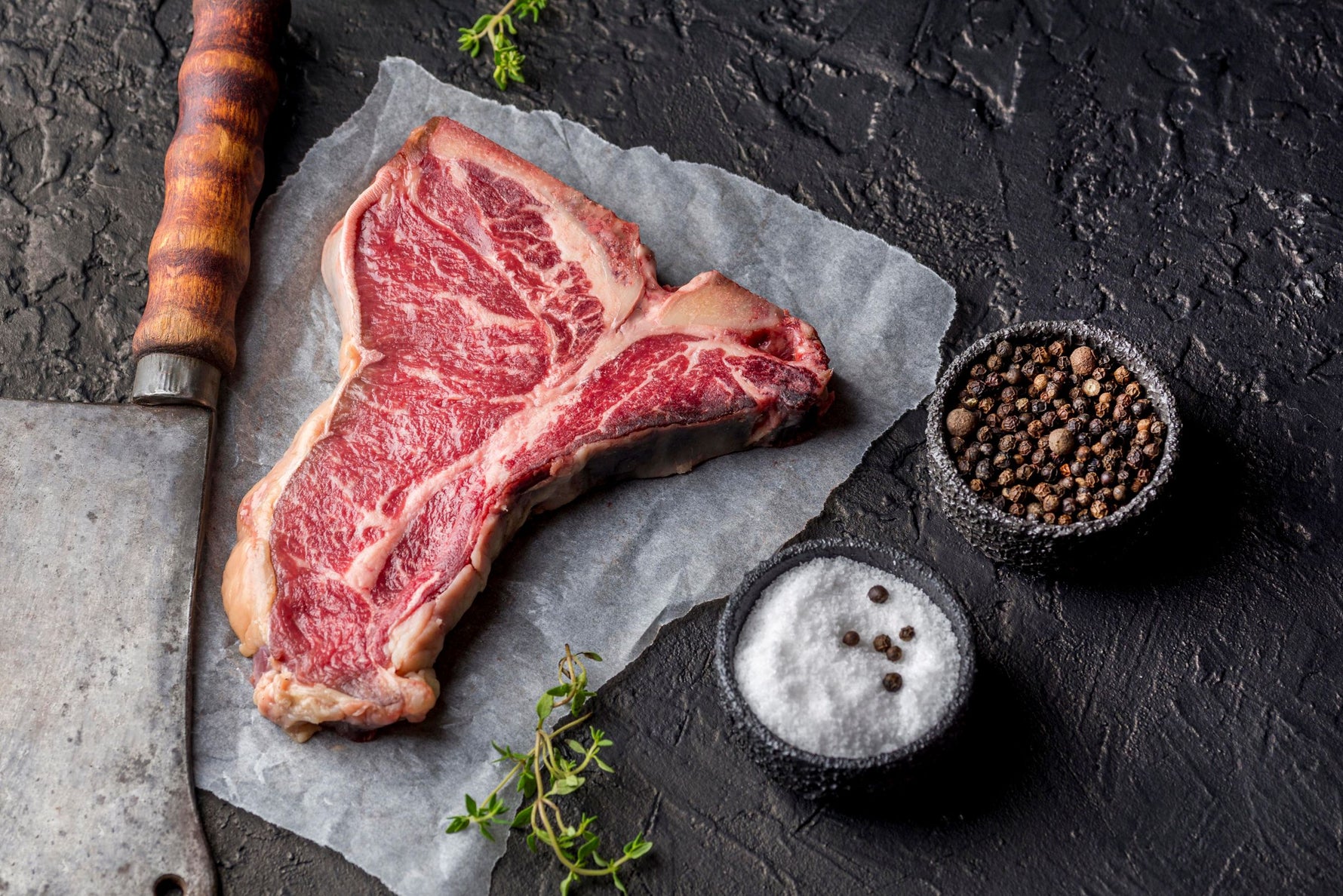 Raw Dry-Aged T-Bone Steak from Irish farmed beef, aged for 28+ days, handcut by butchers on a wooden platter  on parchment paper with salt, pepper, and a knife on a dark surface