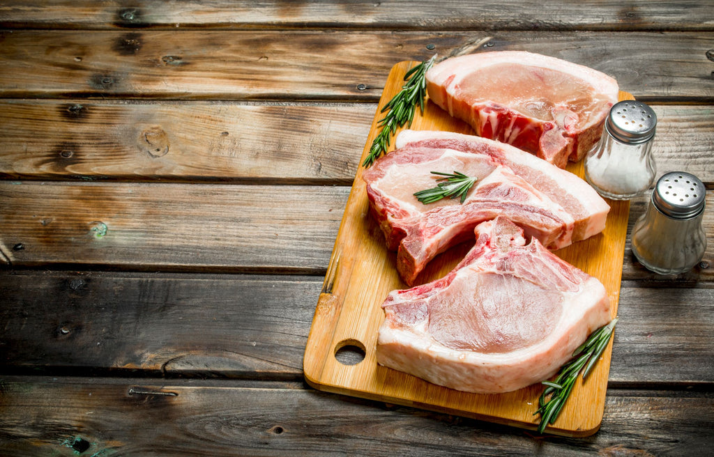 Herterich's Raw pork chops with bone in on a wooden cutting board with rosemary and salt shakers on a rustic wooden surface.