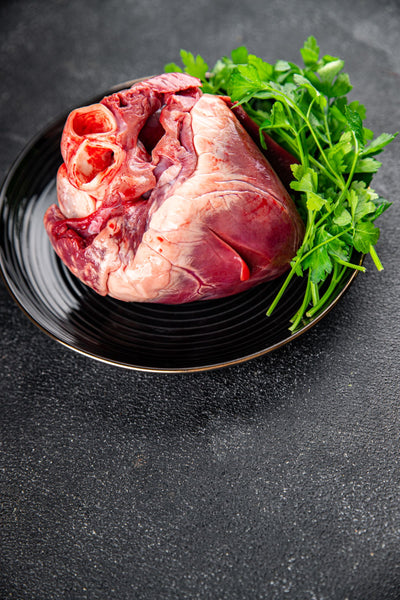 Raw beef heart on a black plate with green herbs on a dark surface