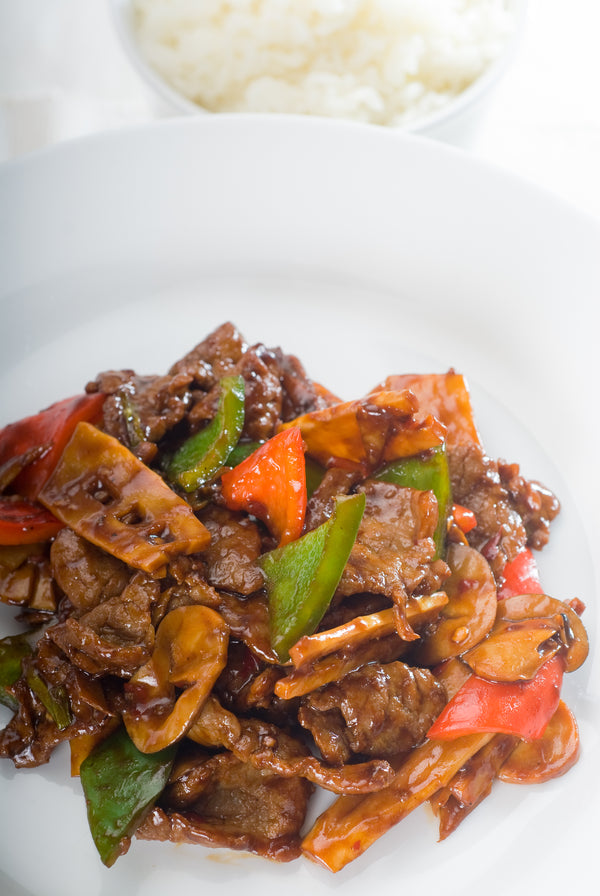 Herterich's Cooked Beef Stir Fry with beef slices, green and red peppers, and other vegetables in a dark sauce, served on a white plate with rice.