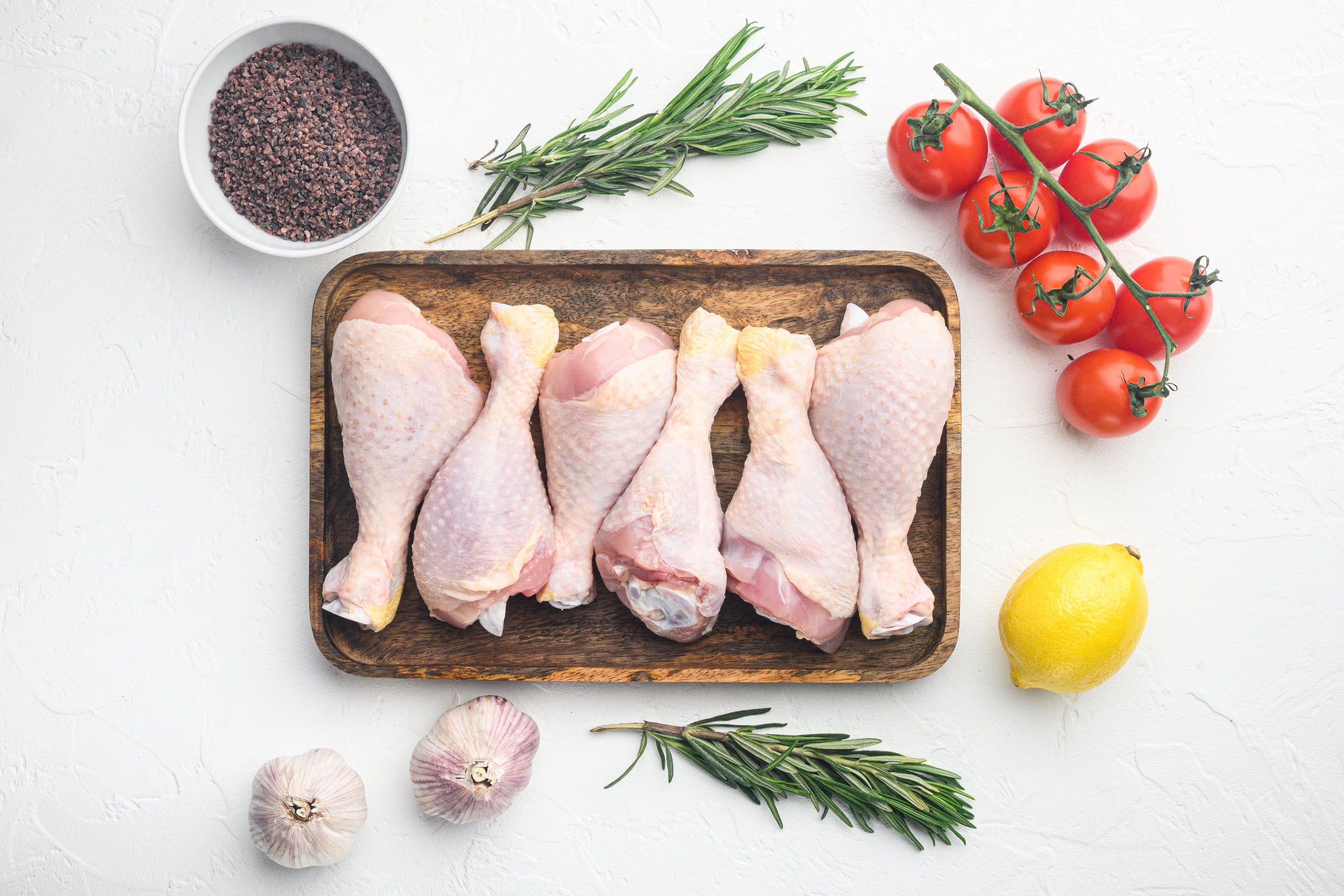 Raw 100% Irish Herterich's Chicken Drumsticks on a wooden tray, with fresh rosemary, cherry tomatoes, lemon, and garlic on a white background.