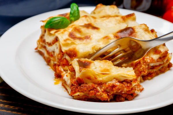 Slice of Herterich's lasagna on a white plate with a fork, garnished with basil.