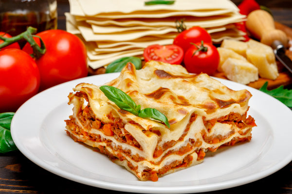 Slice of lasagna on a white plate with tomatoes and pasta in the background