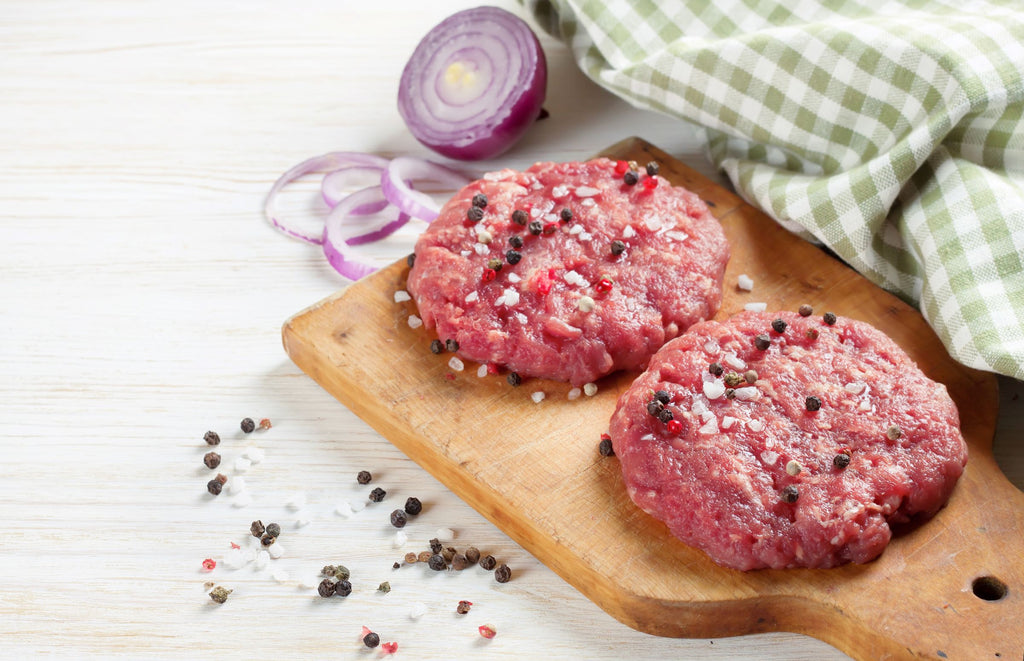 Two raw protein burger patties on a wooden board with onions and spices on a light background
