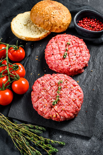 Two raw, hand-shaped 100% Lean Irish Beef Steak Burgers (140g each) with seasoning, ready to cook, on a dark board.