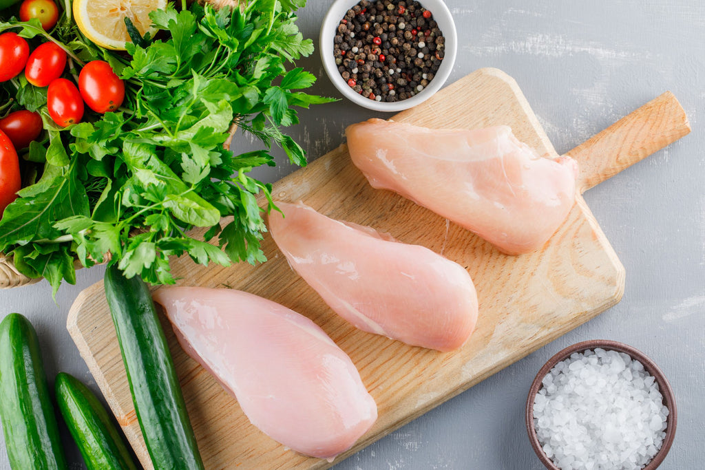 Raw chicken breasts on a wooden cutting board with vegetables and spices on a gray surface